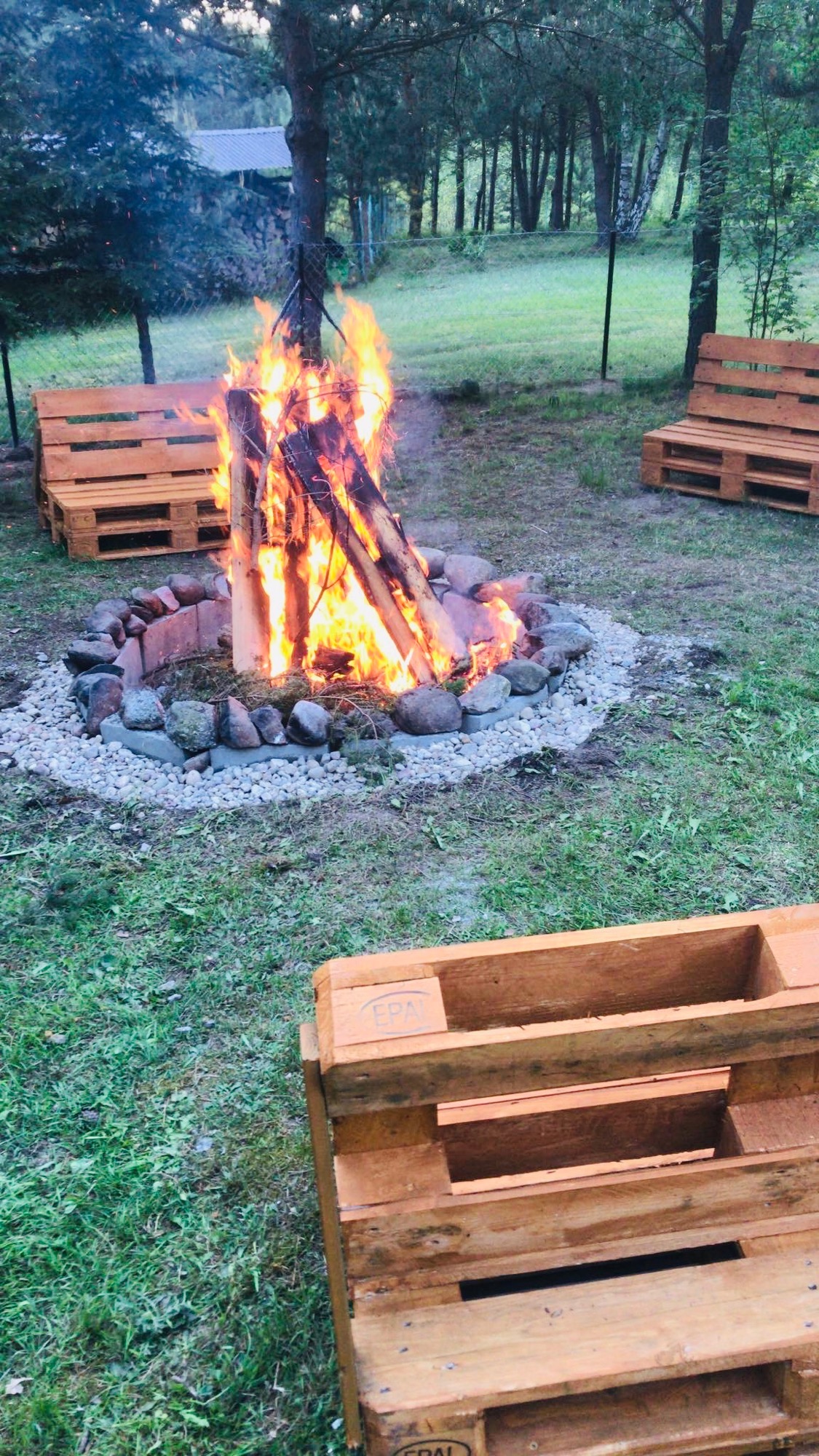 Ognisko otoczone kamieniami i żwirem, w tle dwie drewniane ławki z palet na trawie, w pierwszym planie paleta.