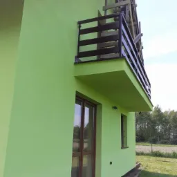 Dom z jasnozieloną elewacją, brązowymi ramami okien i drzwi oraz balkonem z drewnianą balustradą, w tle drzewa.