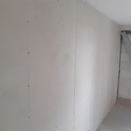 Ściana wewnętrzna wykończona płytami gipsowo-kartonowymi, widoczne wkręty mocujące, surowy stan przed szpachlowaniem, z fragmentem ściany z betonu komórkowego w tle.