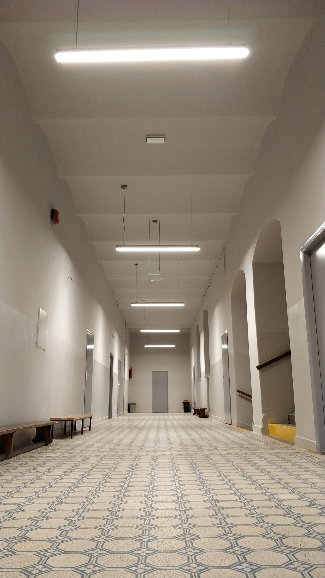 Długi korytarz oświetlony liniowymi lampami LED, z wzorzystą podłogą, ławkami po bokach i widocznymi drzwiami oraz elementami systemu przeciwpożarowego.