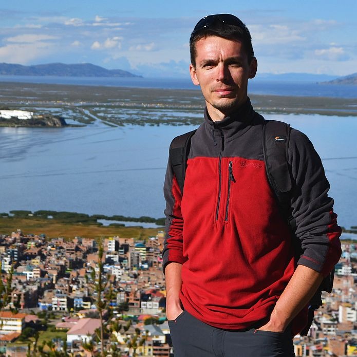 Mężczyzna w czerwono-szarej bluzie polarowej z plecakiem na tle panoramy miasta i jeziora z górami w oddali.