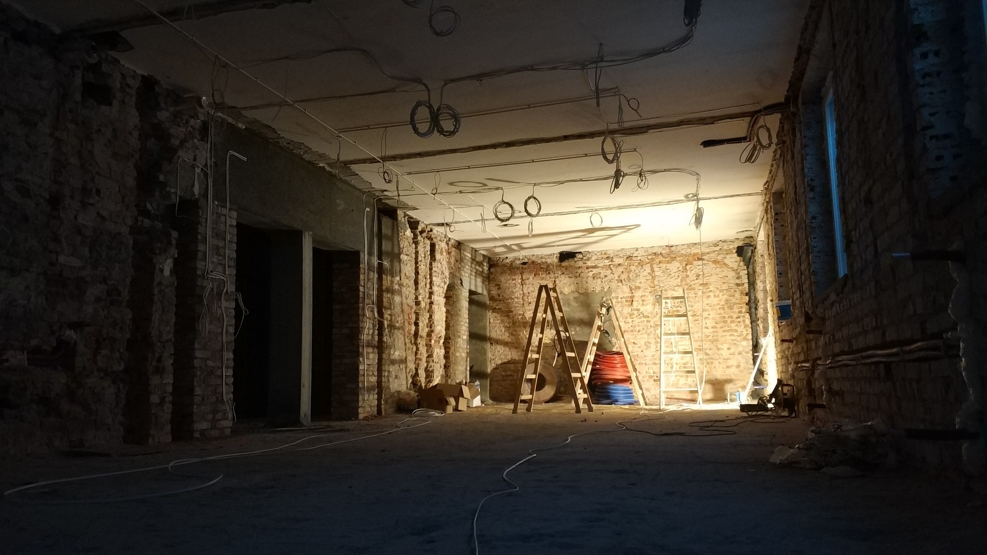 Wnętrze w trakcie remontu z odsłoniętą cegłą, widoczne przewody elektryczne pod sufitem, drabiny opierające się o ścianę oraz zwinięte rury w kolorach czerwonym i niebieskim.