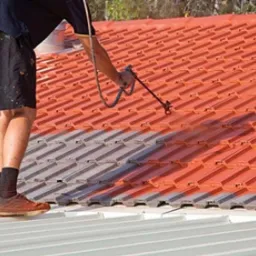 Pracownik w krótkich spodenkach i brązowych butach maluje czerwoną dachówkę pistoletem natryskowym, widoczny fragment pomalowanego dachu i wiadro z farbą.