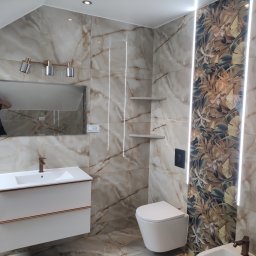 Artur Olejarnik - Wykończona łazienka w beżowych płytkach imitujących marmur, z akcentem w postaci ściany z motywem roślinnym podświetlonej ledami. Widoczna umywalka, toaleta i lustro.