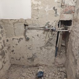 Artur Olejarnik - Surowa ściana łazienki w trakcie remontu z odsłoniętą instalacją wodną i gruzem na podłodze. Widoczne rury i bateria z mieszaczem, fragment ściany pomalowany na biało.