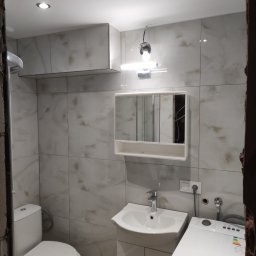 Artur Olejarnik - Wykończona łazienka z marmurowymi płytkami, umywalką z szafką, toaletą i pralką. Widoczne oświetlenie sufitowe i lustro z szafką. Częściowo widoczne futryny.