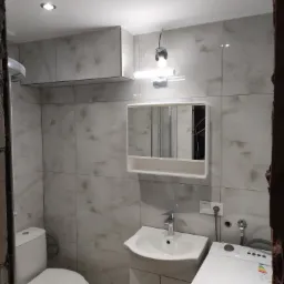 Wykończona łazienka z marmurowymi płytkami, umywalką z szafką, toaletą i pralką. Widoczne oświetlenie sufitowe i lustro z szafką. Częściowo widoczne futryny.