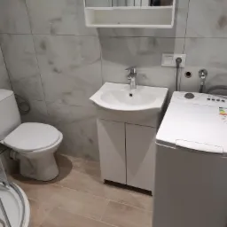 Nowoczesna łazienka po remoncie: biała umywalka z szafką, toaleta, pralka i kabina prysznicowa. Jasne płytki imitujące marmur na ścianach i podłodze.