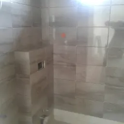 Ściana łazienki wyłożona szarymi, błyszczącymi płytkami z widocznym otworem na rurę i puszką elektryczną, fuga biała, widoczne krzyżyki dystansowe.