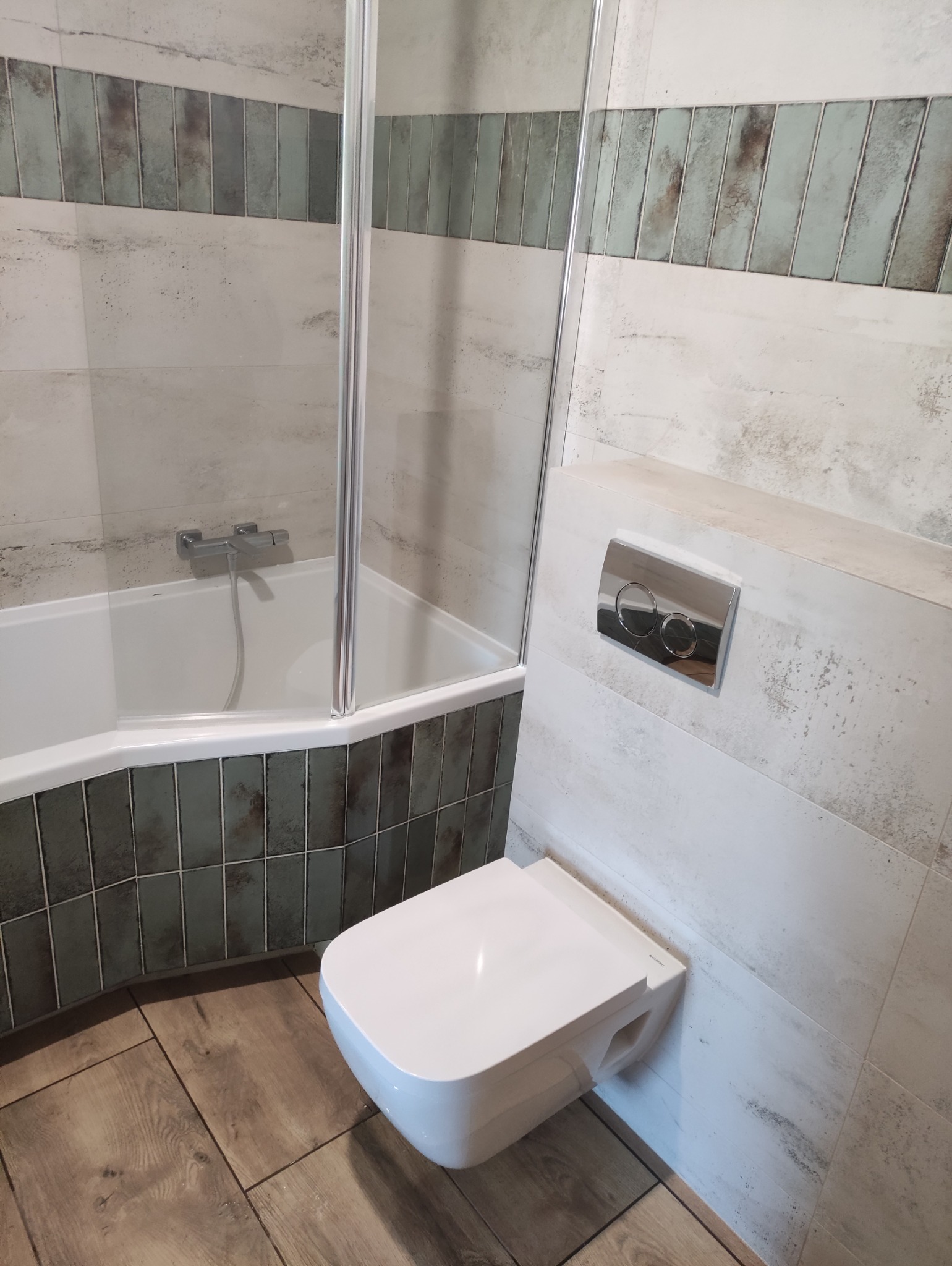 Nowoczesna łazienka po remoncie: podwieszana toaleta z chromowanym przyciskiem, kabina prysznicowa i płytki imitujące drewno na podłodze.