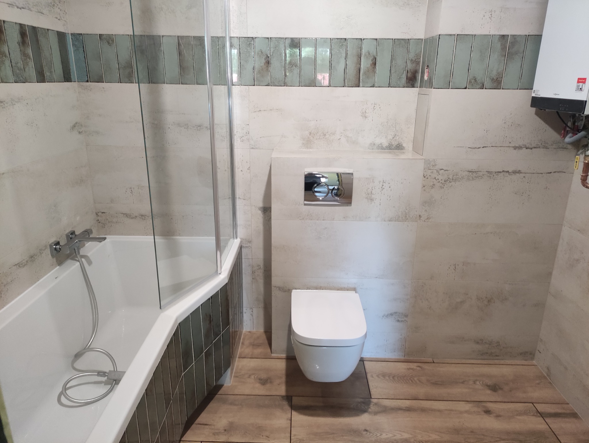 Wykończona łazienka z beżowymi płytkami imitującymi kamień, drewnopodobną podłogą i zabudowaną toaletą. Widoczny bojler i fragment kabiny prysznicowej.