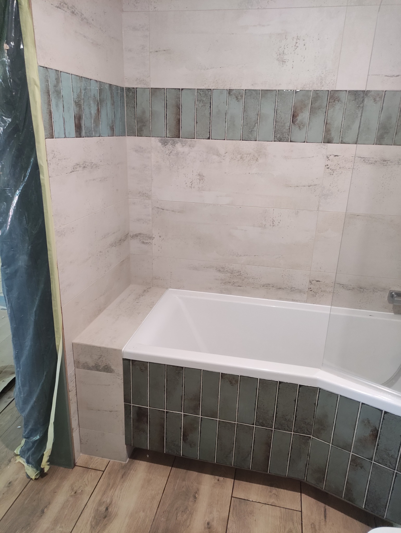 Wykończona łazienka z wanną obłożoną płytkami w odcieniach szarości i bieli, z akcentem w postaci dekoracyjnych płytek w kolorze morskim. Widoczne drewnopodobne panele na podłodze.