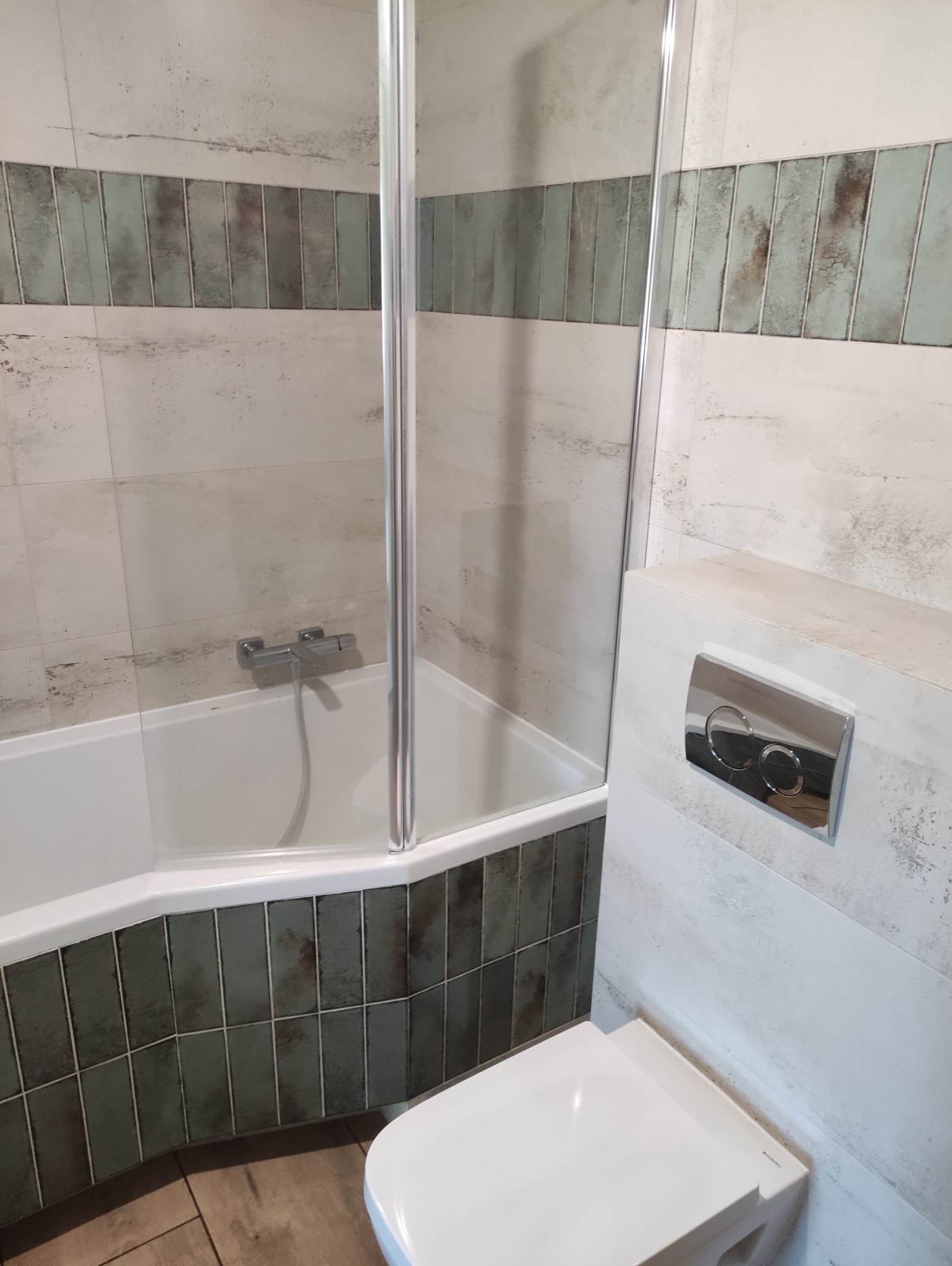 Wykończona łazienka z wanną za szklaną ścianką, toaleta podwieszana i płytki w odcieniach bieli i zieleni. Widok na fragment wnętrza łazienki po remoncie.