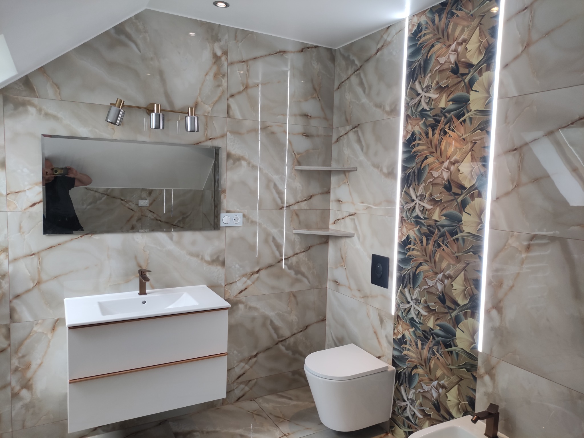 Wykończona łazienka w beżowych płytkach imitujących marmur, z akcentem w postaci ściany z motywem roślinnym podświetlonej ledami. Widoczna umywalka, toaleta i lustro.