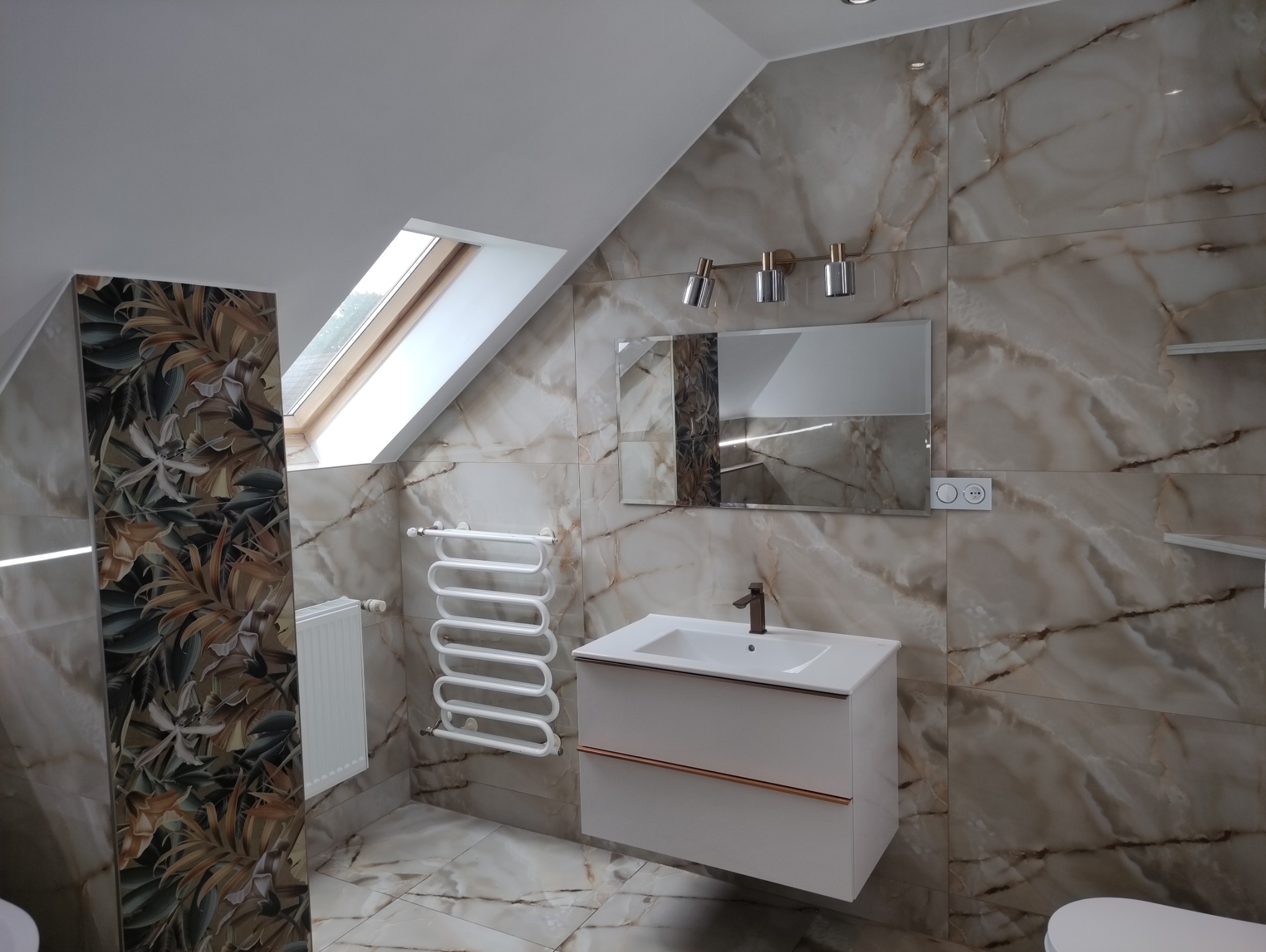 Wykończona łazienka na poddaszu z beżowymi płytkami imitującymi marmur, białą szafką z umywalką i grzejnikiem drabinkowym. Widoczne okno dachowe i oświetlenie.