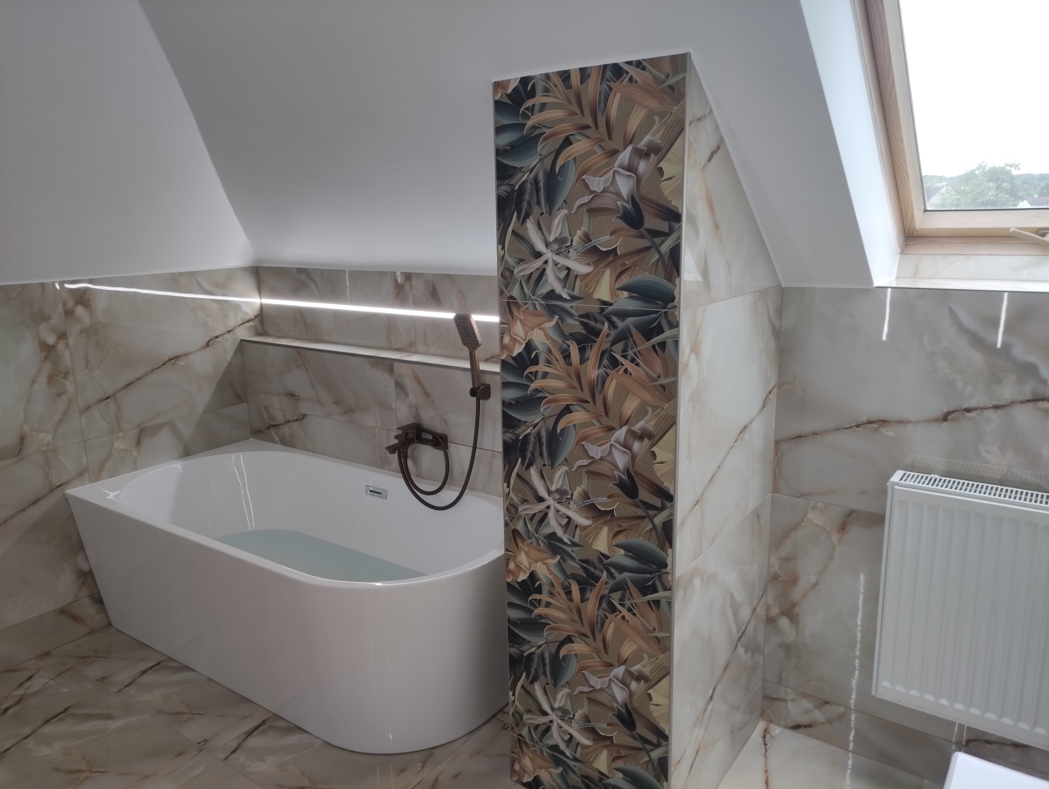 Wykończona łazienka na poddaszu z białą wanną, płytkami imitującymi marmur i dekoracyjnym panelem z motywem roślinnym przy wannie. Okno dachowe w tle.