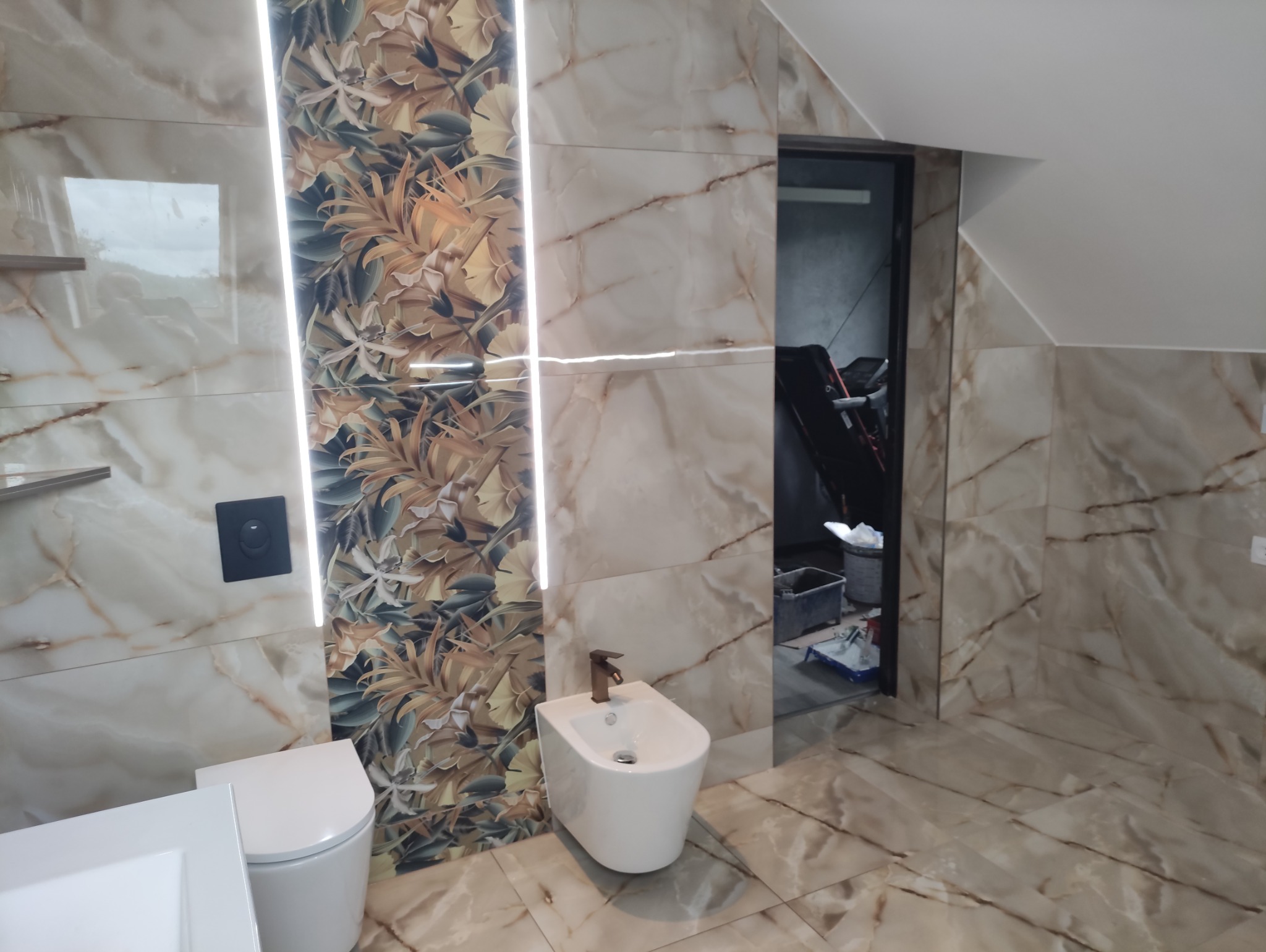 Wykończona łazienka na poddaszu z marmurowymi płytkami i motywem florystycznym, białe sanitariaty, widoczne narzędzia malarskie w tle.