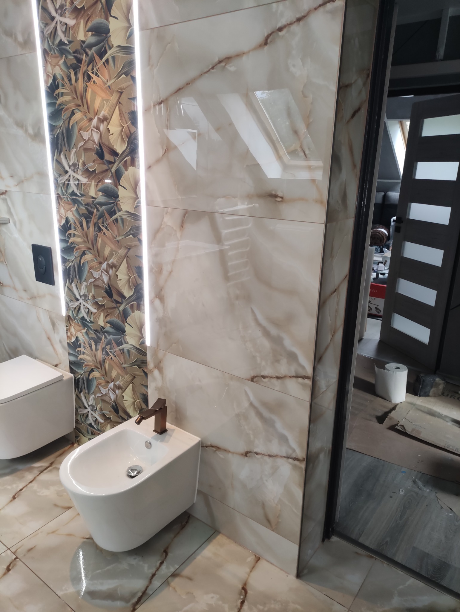 Wykończona łazienka z beżowymi płytkami imitującymi marmur, dekoracyjną ścianą z motywem roślinnym i podświetleniem LED. Widoczne bidet i toaleta.