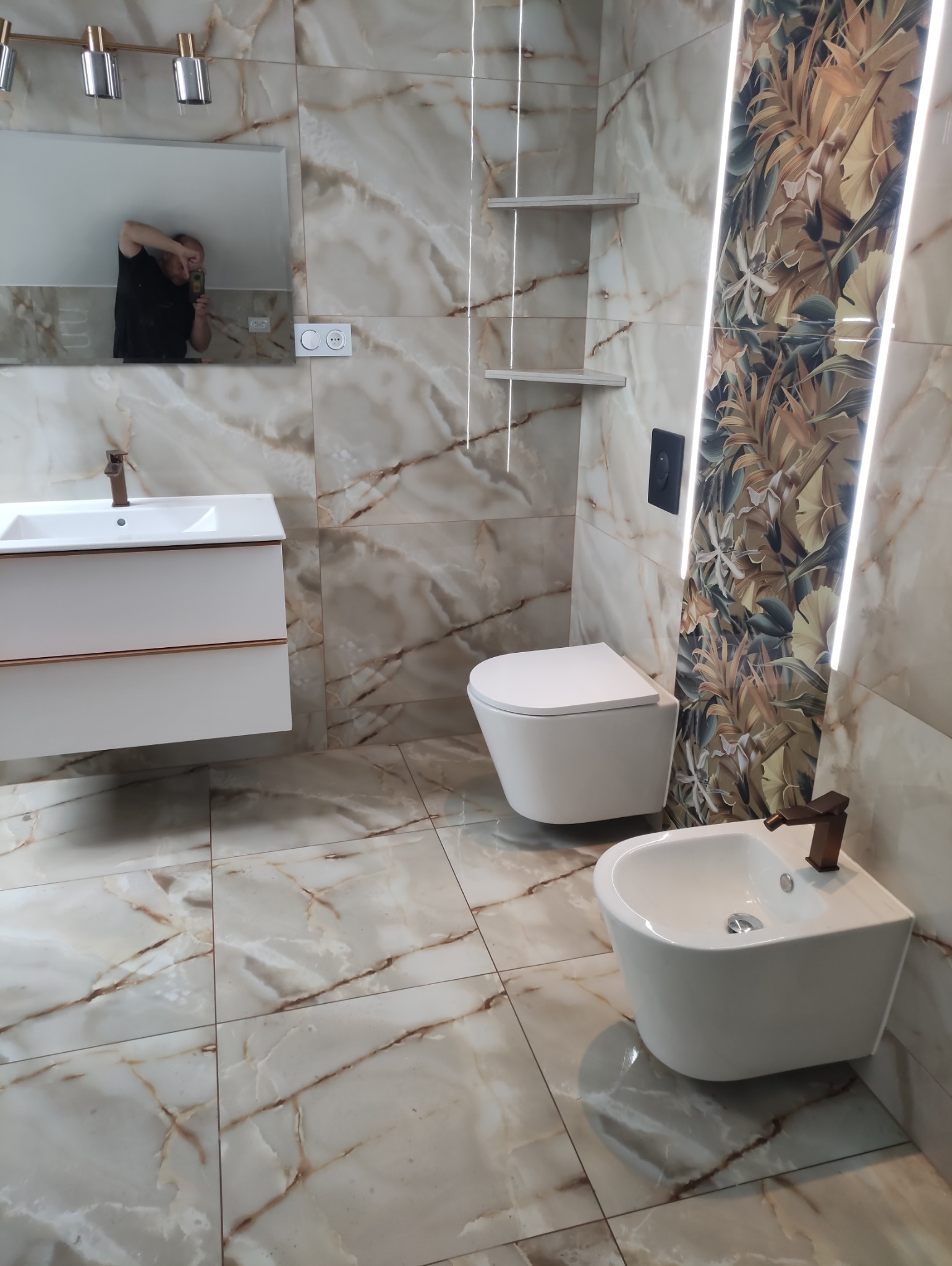 Wykończona łazienka z płytkami imitującymi marmur, podświetleniem LED, wiszącą toaletą i bidetem. W lustrze odbija się fotograf.