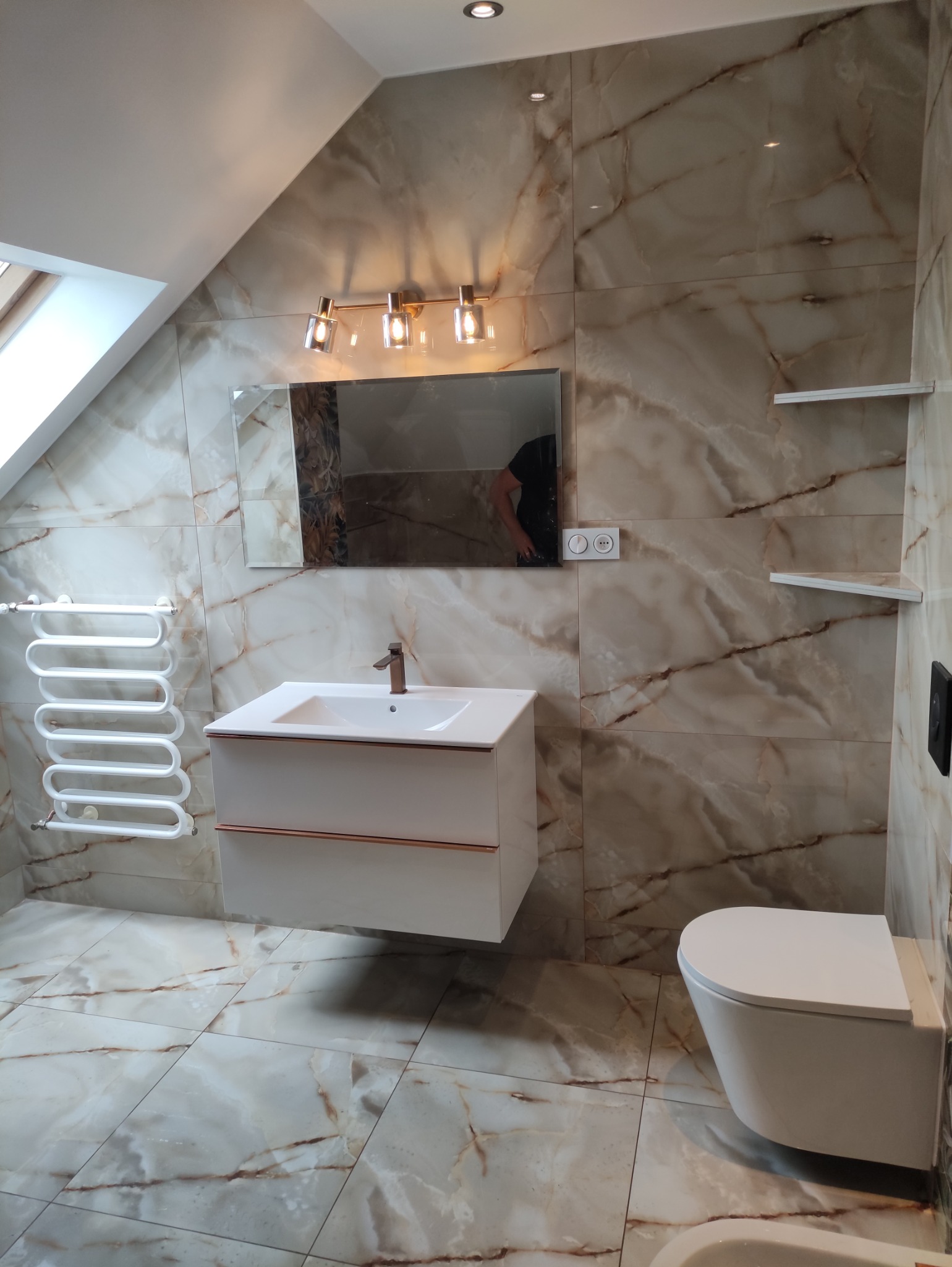 Wykończona łazienka na poddaszu z marmurowymi płytkami, wiszącą szafką z umywalką, grzejnikiem drabinkowym i toaletą. Uwagę zwraca skośny sufit z oknem.