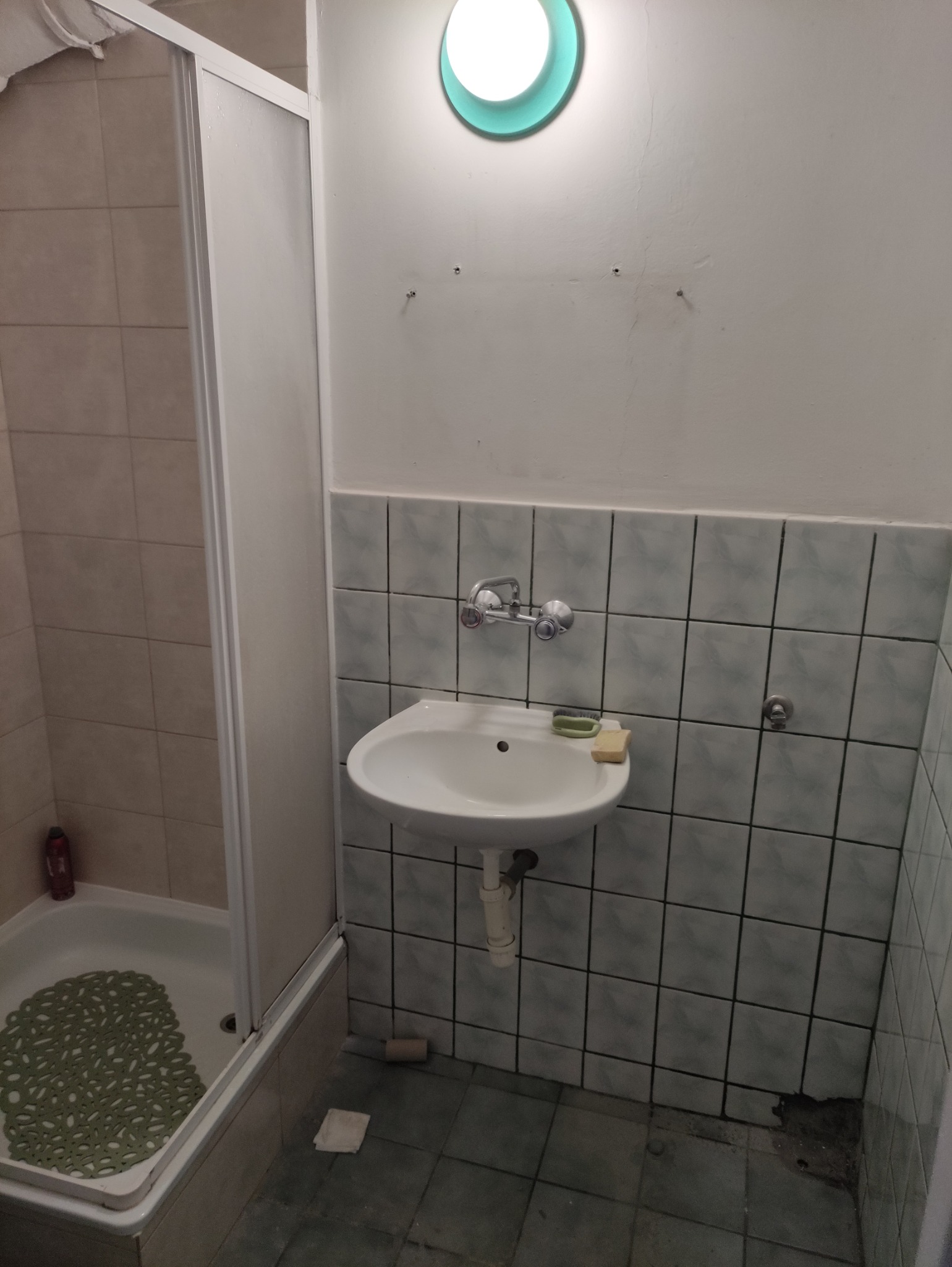 Umywalka z baterią i mydłem na tle wyłożonej płytkami ściany w łazience z kabiną prysznicową. Widoczne ślady użytkowania i niedociągnięcia.