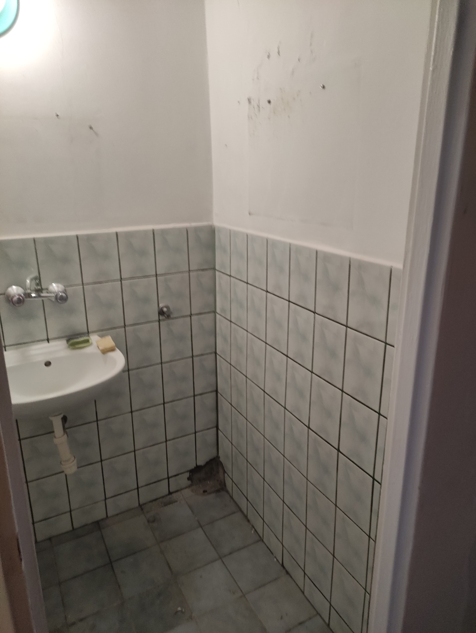 Wnętrze małej łazienki z umywalką i kafelkami w odcieniach szarości i zieleni. Widoczne ślady użytkowania na ścianach i podłodze, co sugeruje potrzebę remontu.