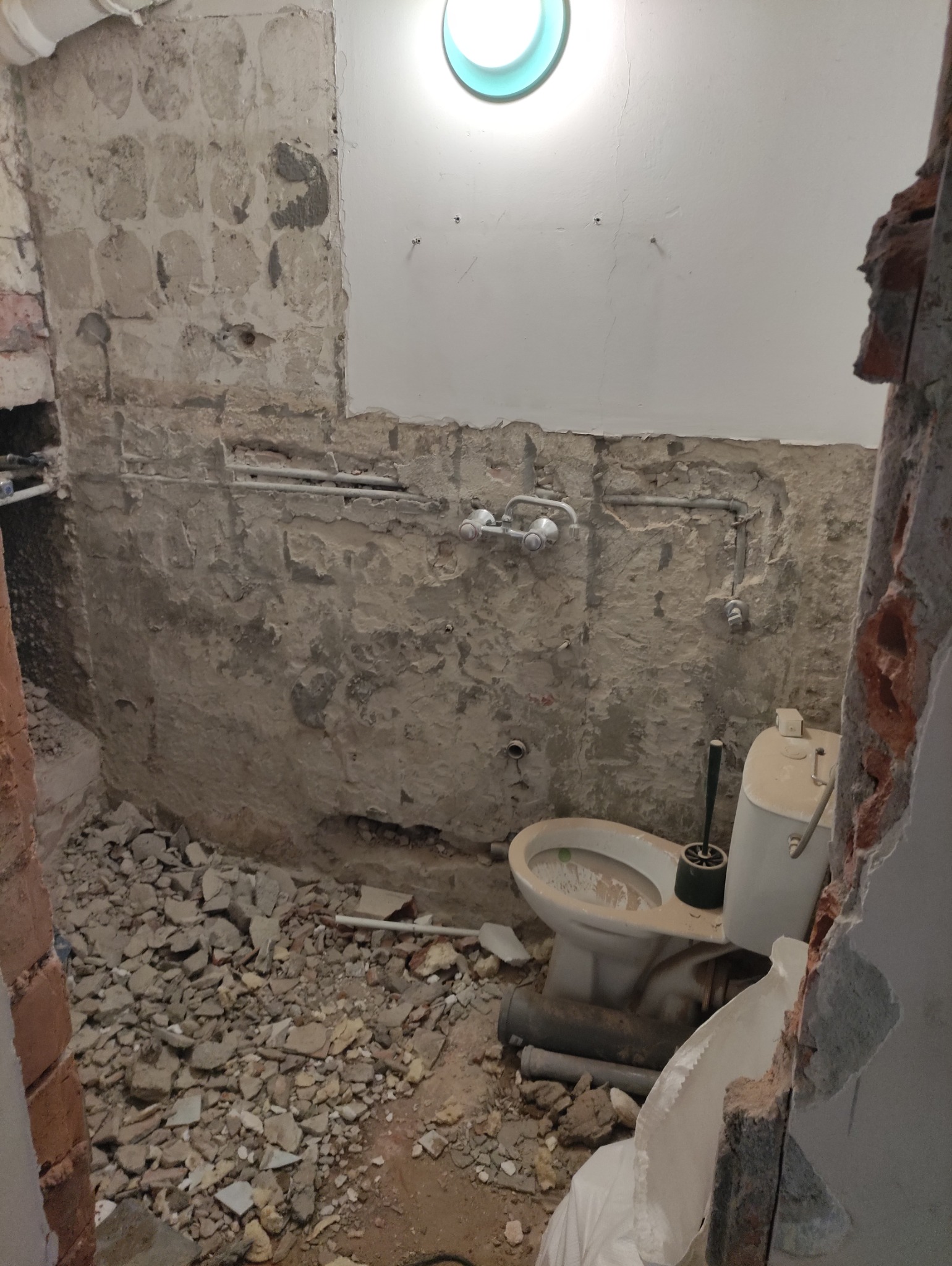 Łazienka w trakcie demontażu: gruz na podłodze, odsłonięte rury, stara toaleta. Ściany częściowo skute, widoczne ślady po płytkach. Etap przygotowawczy remontu.