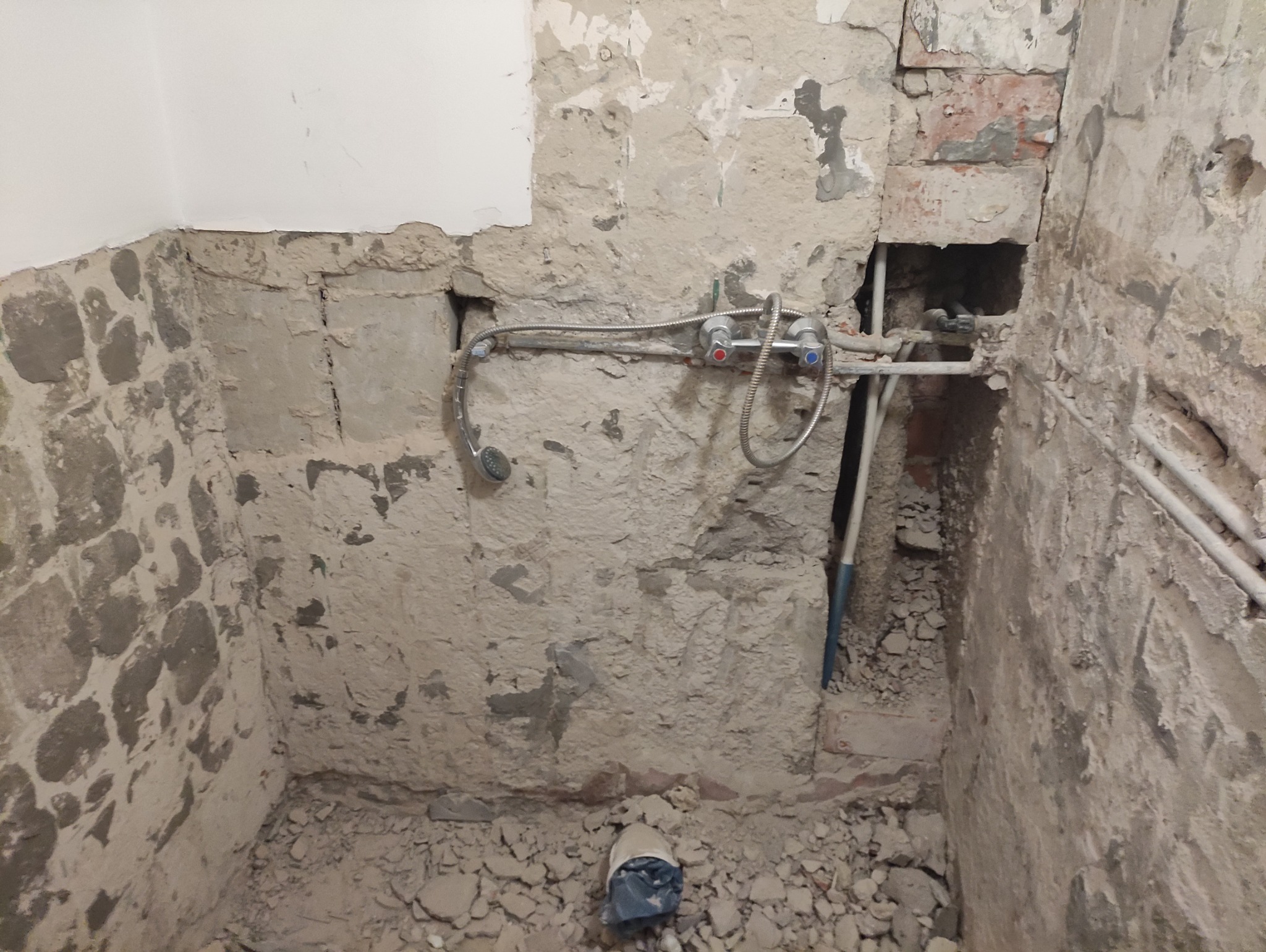 Surowa ściana łazienki w trakcie remontu z odsłoniętą instalacją wodną i gruzem na podłodze. Widoczne rury i bateria z mieszaczem, fragment ściany pomalowany na biało.