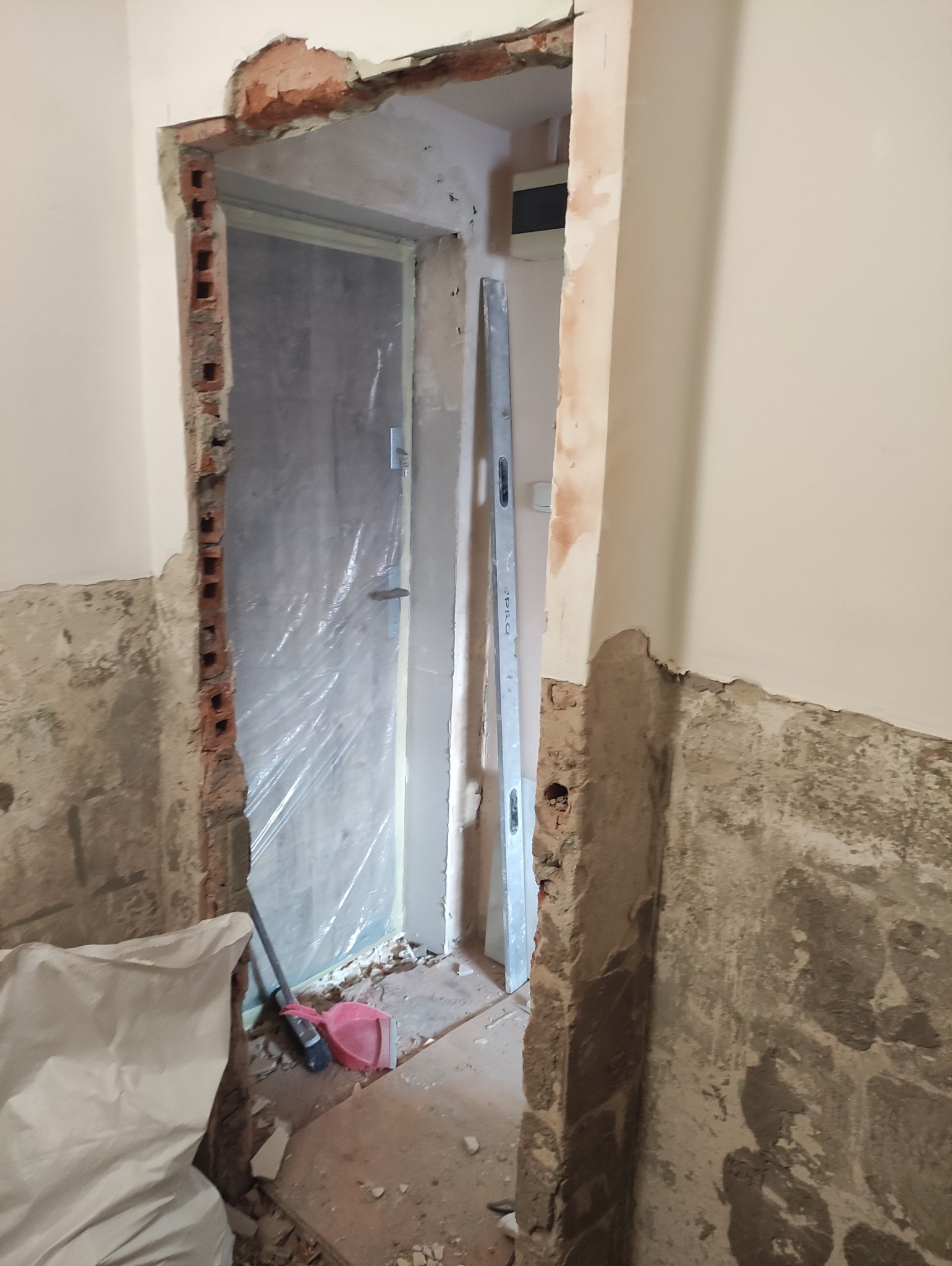 Demontaż ościeżnicy drzwiowej w trakcie remontu. Widoczne gruz, poziomica, resztki muru i folia ochronna na drzwiach. Fragment ściany w stanie surowym.
