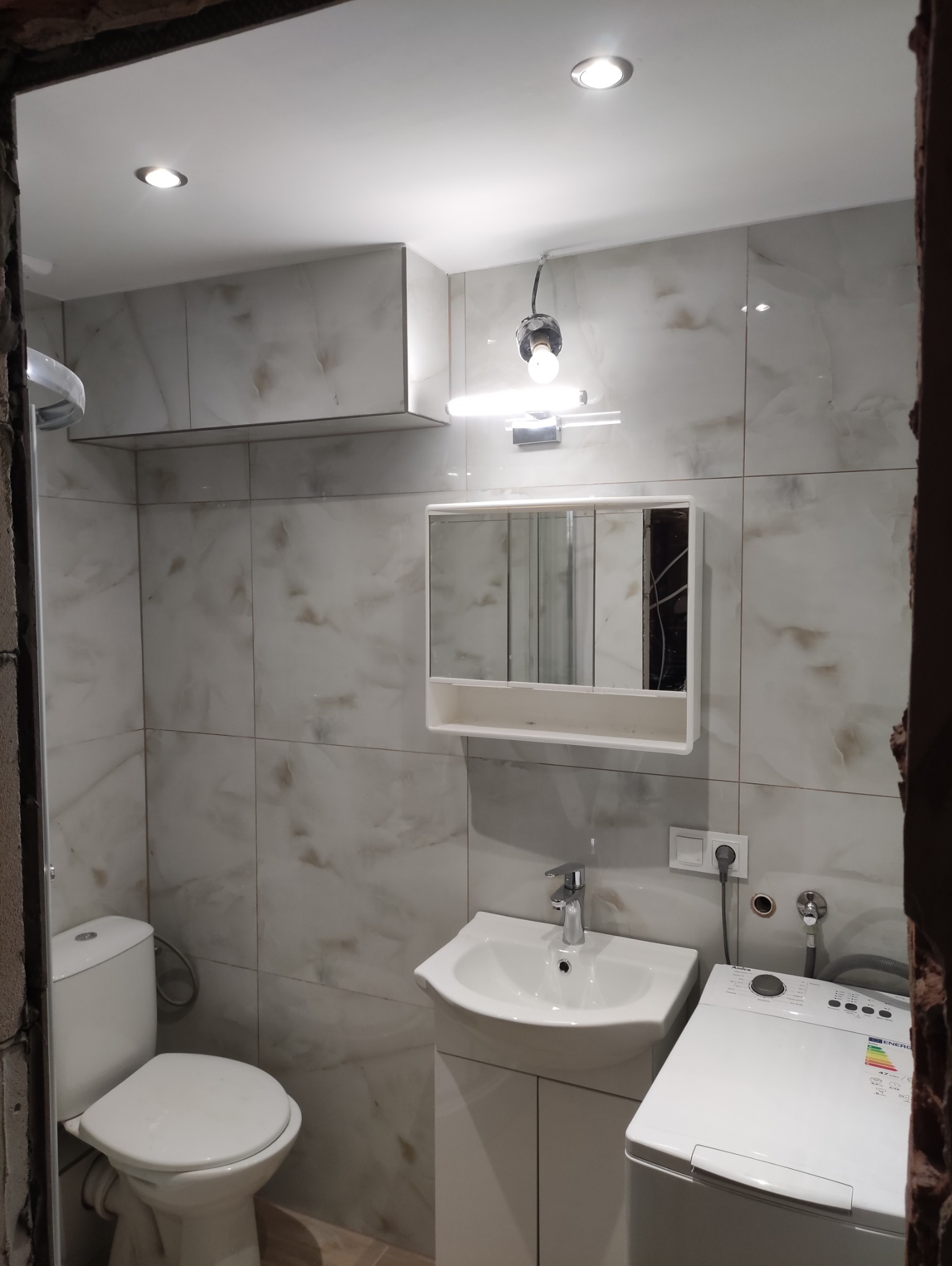 Wykończona łazienka z marmurowymi płytkami, umywalką z szafką, toaletą i pralką. Widoczne oświetlenie sufitowe i lustro z szafką. Częściowo widoczne futryny.