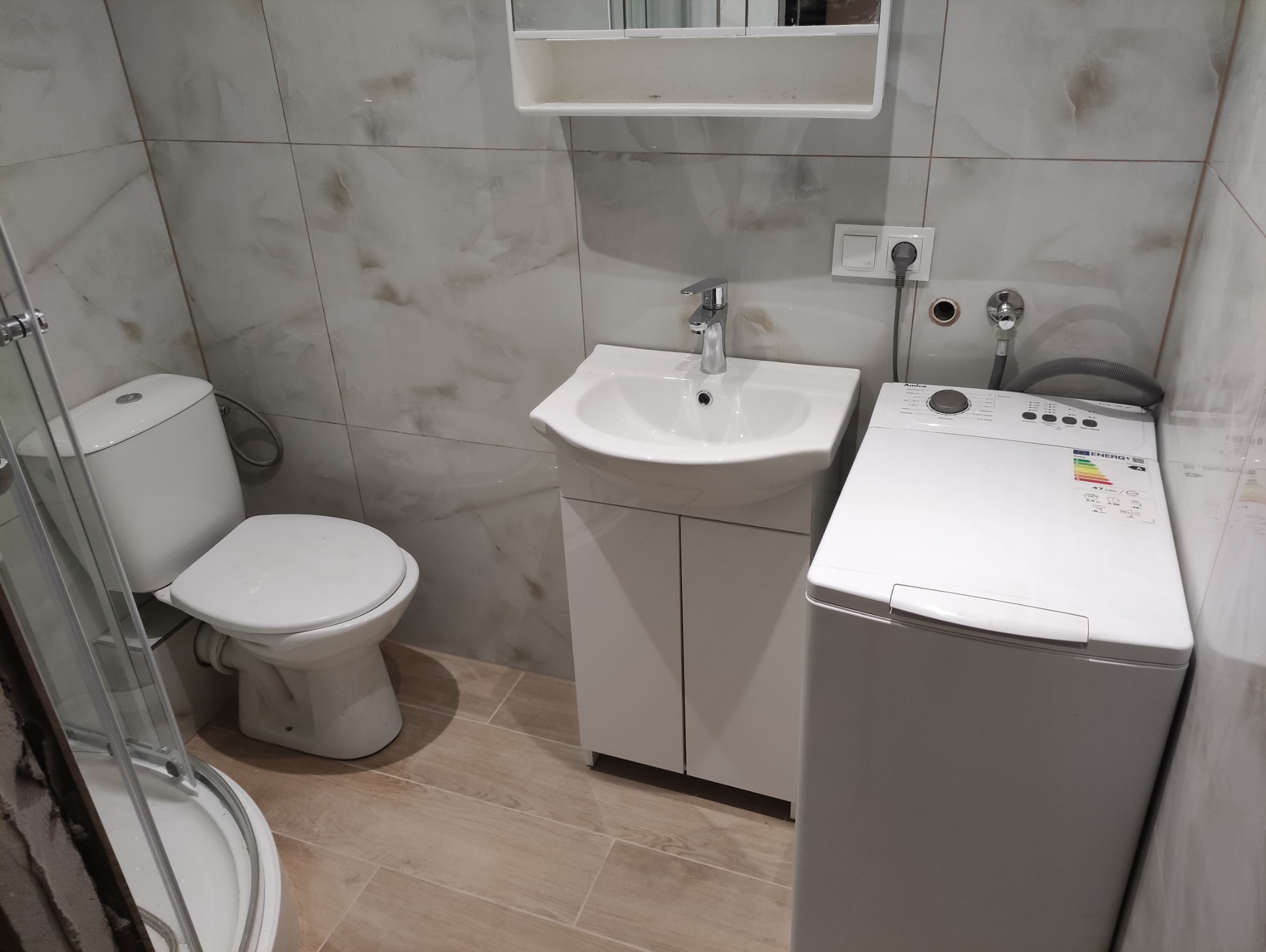 Nowoczesna łazienka po remoncie: biała umywalka z szafką, toaleta, pralka i kabina prysznicowa. Jasne płytki imitujące marmur na ścianach i podłodze.