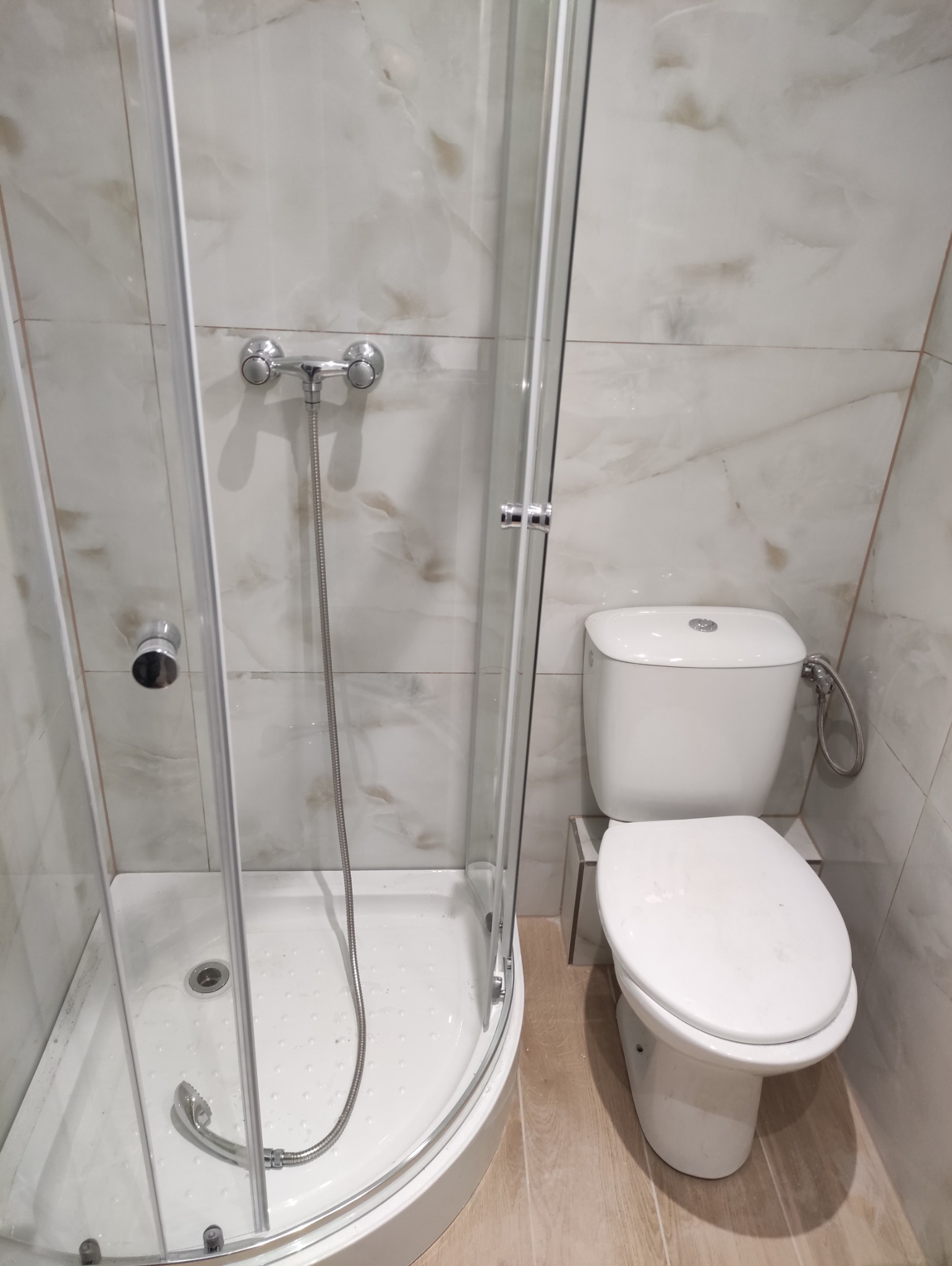 Wykończona łazienka: kabina prysznicowa z brodzikiem i toaleta w jasnych, marmurkowych płytkach. Widok na armaturę i detale wykończenia.