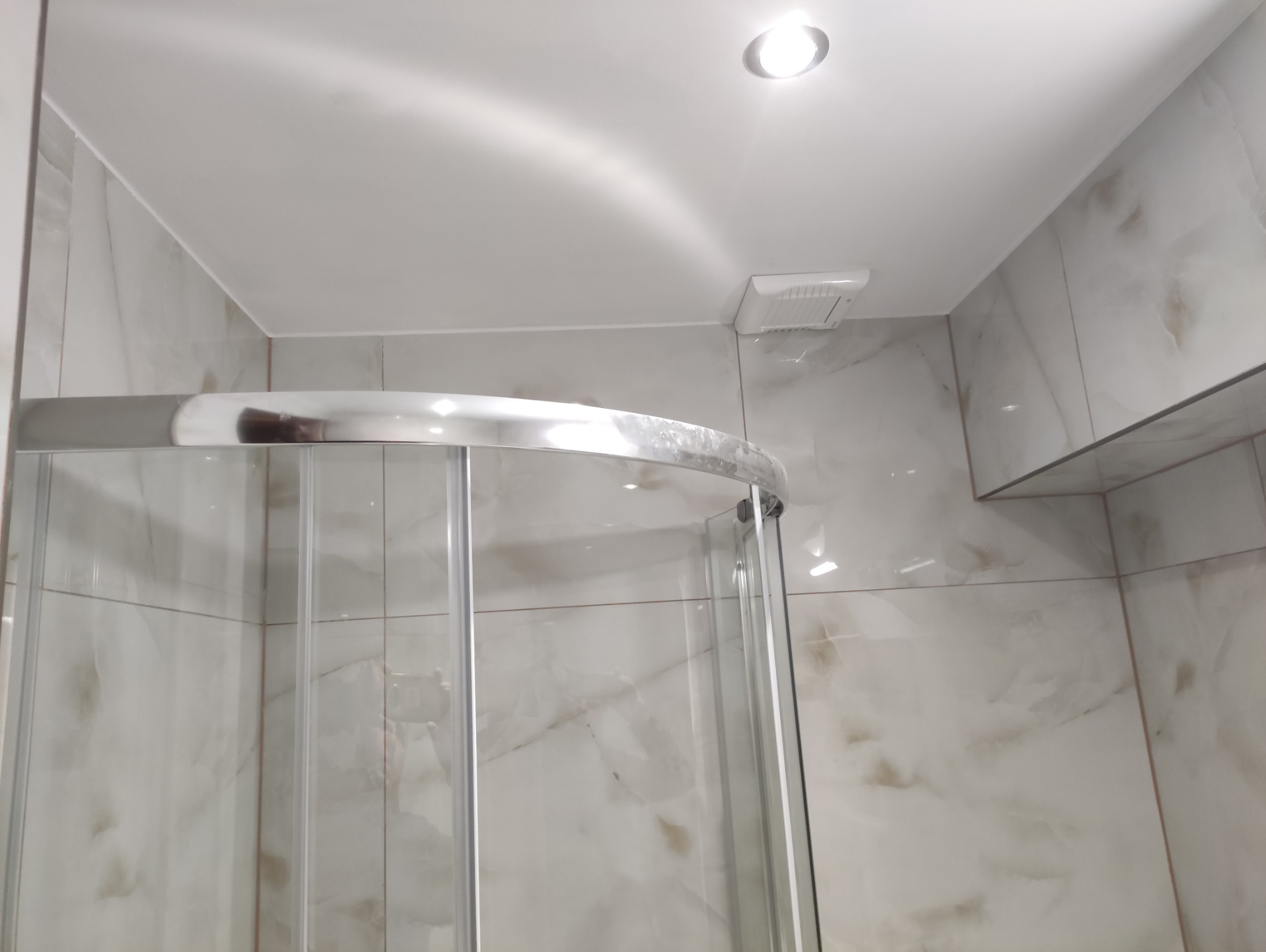 Wykończona łazienka z kabiną prysznicową z chromowanymi profilami i marmurowymi płytkami. Widoczny wentylator i oświetlenie sufitowe.