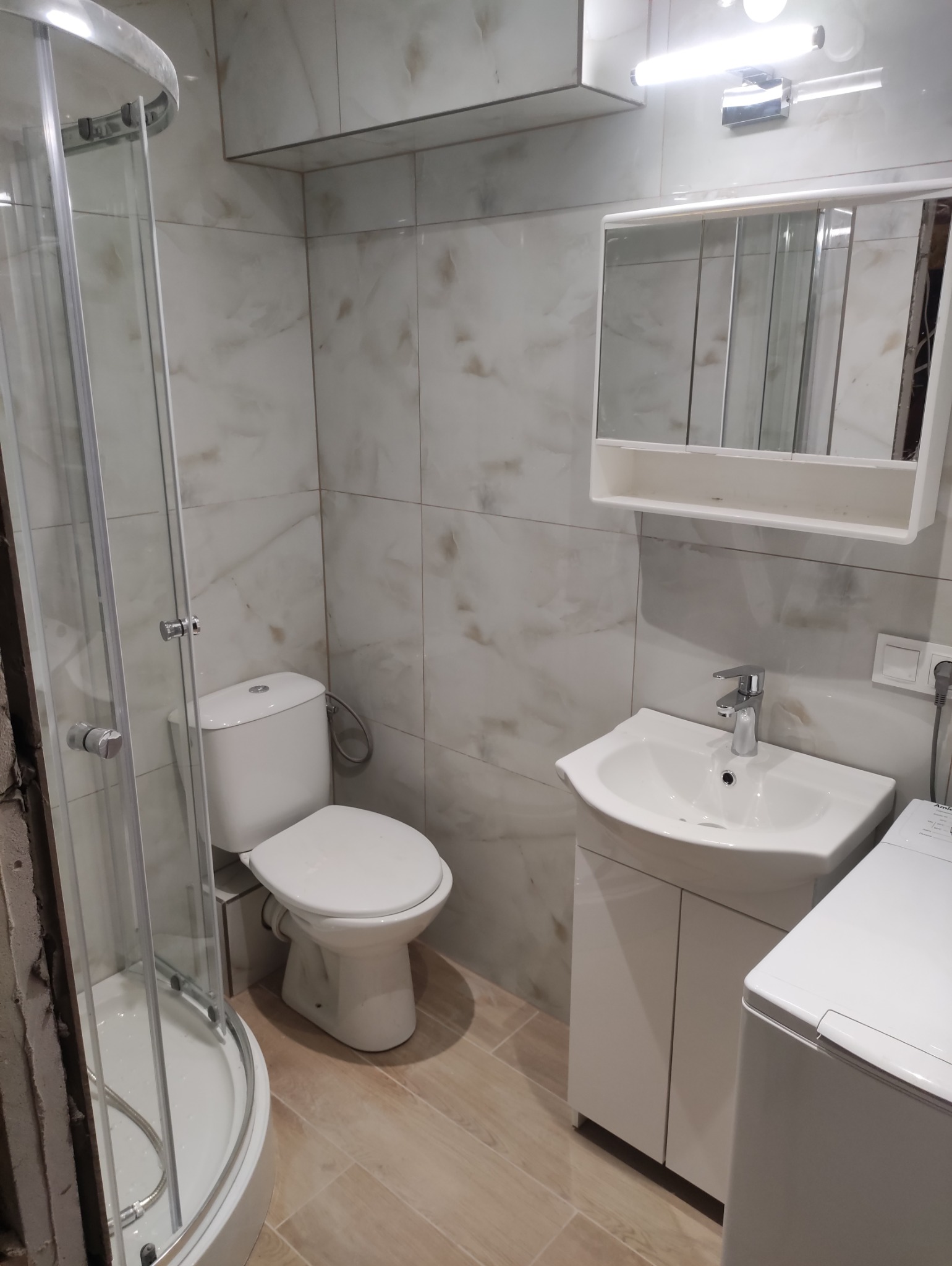 Wykończona łazienka z kabiną prysznicową, toaletą, umywalką z szafką i pralką. Jasne płytki imitujące marmur na ścianach i podłodze. Nowoczesne oświetlenie.