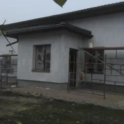 Dom jednorodzinny w trakcie ocieplania elewacji, widoczne rusztowania, okna zabezpieczone folią, świeżo nałożony tynk, pochmurne niebo.