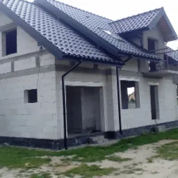 Niedokończony dom jednorodzinny z białych bloczków, z zamontowaną czarną dachówką i rynnami, widoczne rusztowanie metalowe, otoczenie trawnik i drzewa.