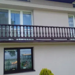 Dwupiętrowy dom z jasną elewacją, brązową balustradą balkonu i oknami z białymi ramami, otoczony zielenią.