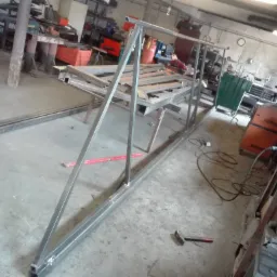 Metalowa konstrukcja z profili kwadratowych w warsztacie, widoczne ślady spawania, młotek na podłodze, spawarka w tle.