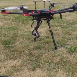 Profesjonalny dron z zamontowaną kamerą, stojący na trójnogu na łące, w tle zabudowania wiejskie pod pochmurnym niebem. W dolnej części kadru widoczny tablet.