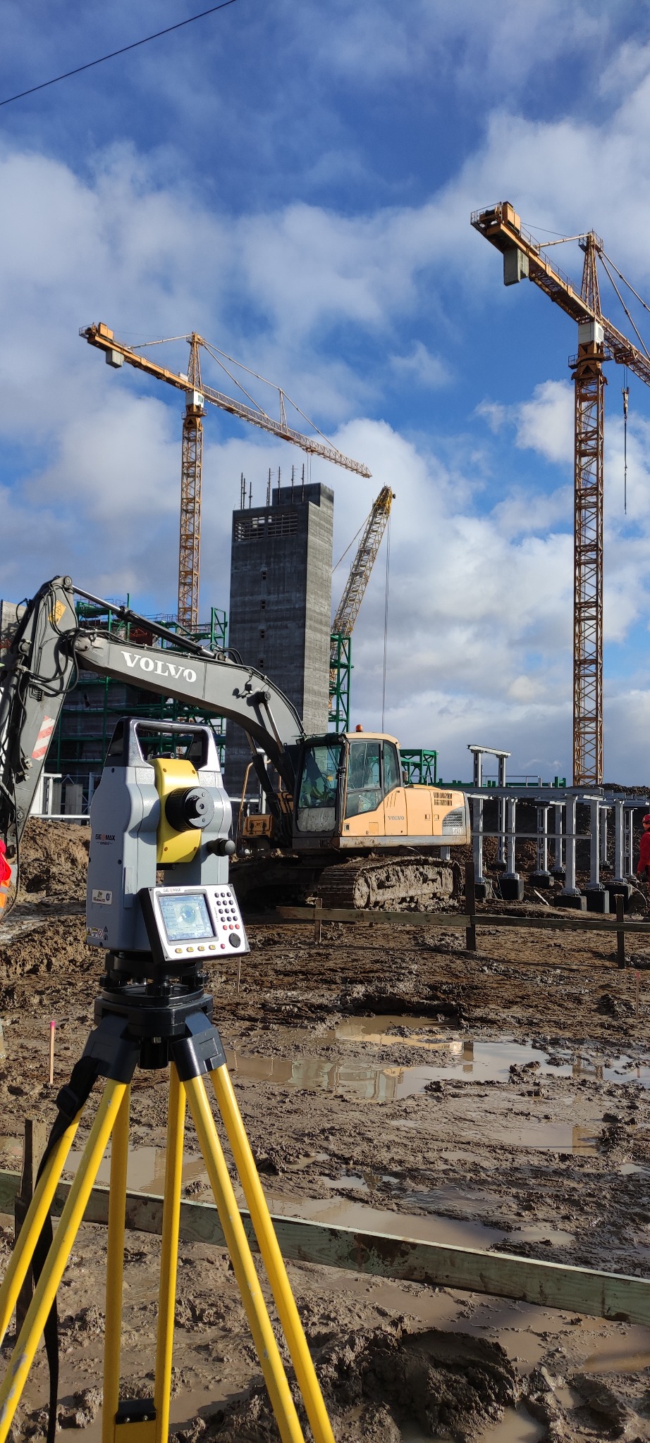 Żółty teodolit na trójnogu na błotnistym placu budowy z koparką Volvo i trzema żurawiami w tle, przy pochmurnym niebie.