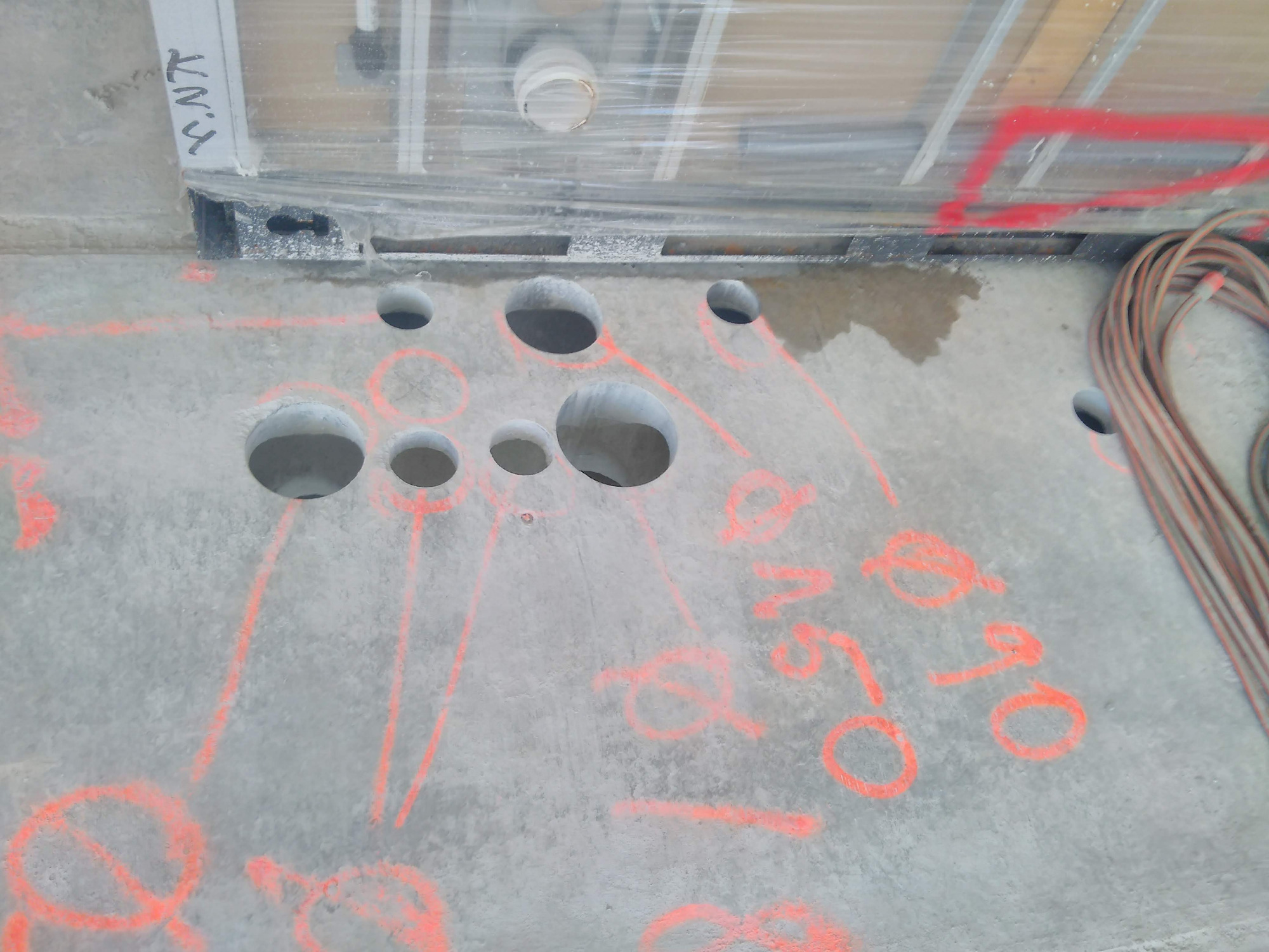 Wiercenie otworów w betonowym fundamencie, pomiar średnicy otworów zaznaczony czerwoną farbą, widoczne przewody elektryczne i fragment instalacji pod stropem.
