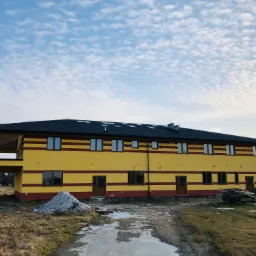 Dwukondygnacyjny budynek z żółtą elewacją w poziome pasy, czarny dach z lukarnami, sterta gruzu przed budynkiem, błotnista droga dojazdowa.