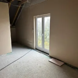 Pok&oacute;j na poddaszu po nałożeniu gładzi gipsowej na ściany, widoczne okno balkonowe, instalacja elektryczna w peszlach i surowe drewniane elementy konstrukcji dachu.