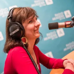 Kobieta uśmiecha się do mikrofonu w studiu radiowym, nosząc słuchawki HD-660 i kolczyk z motywem róży.