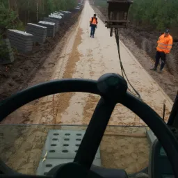 Widok z kabiny maszyny budowlanej na utwardzaną piaszczystą drogę z ułożonymi obok betonowymi płytami, pracownicy w odblaskowych kamizelkach, w tle las.