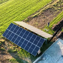Widok z góry na instalację paneli słonecznych na trawiastym terenie, obok wykopany rów, pracownik w oddali i mała koparka JCB na betonowym podłożu