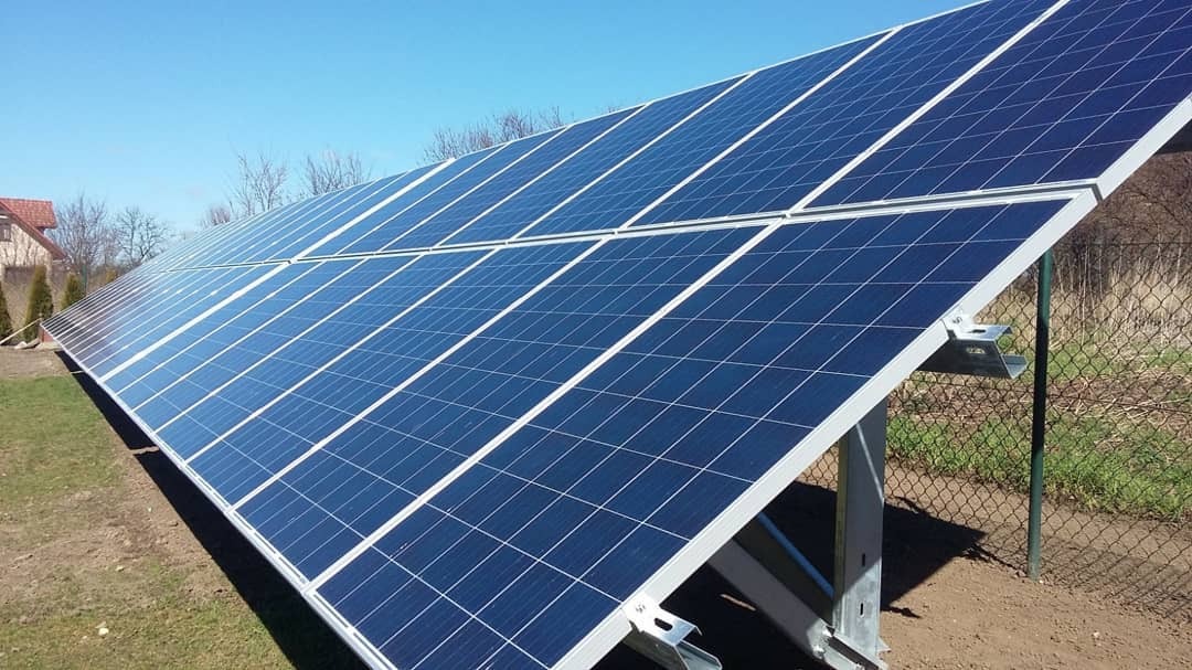 Instalacja paneli słonecznych na gruncie, widoczne metalowe konstrukcje wsporcze, w tle budynek mieszkalny i drzewa, błękitne niebo.