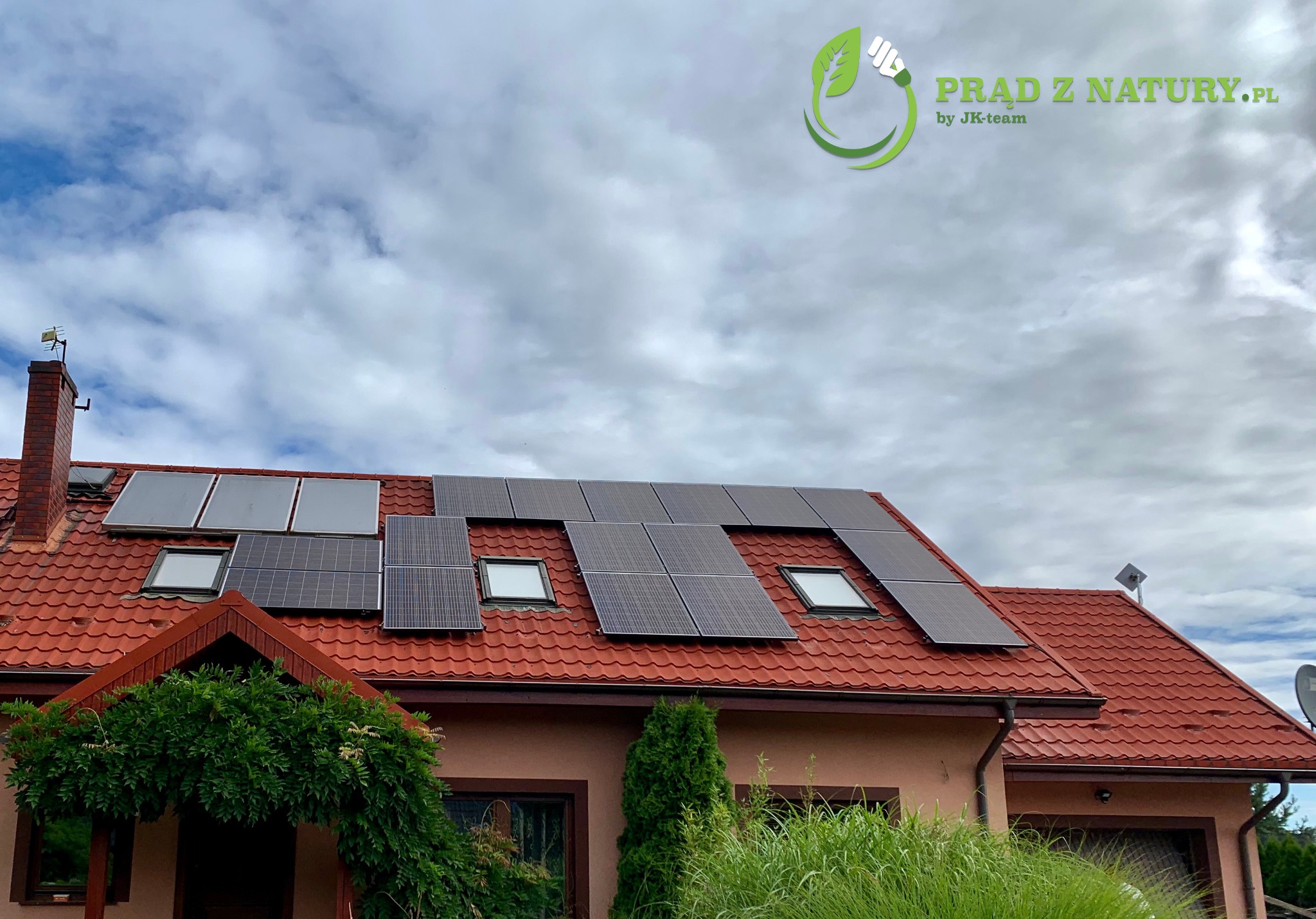 Dom jednorodzinny z czerwoną dachówką i panelami słonecznymi na dachu, widoczne okna dachowe i logo firmy na górze zdjęcia na tle pochmurnego nieba.