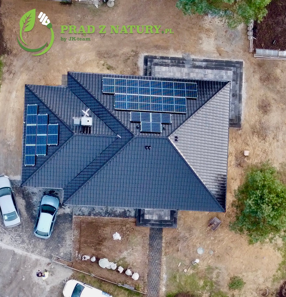 Widok z drona na dom z ciemnym dachem, na którym zamontowano panele słoneczne; widoczne trzy samochody, częściowo wybrukowane podwórko i logo firmy 'Prąd z Natury' w lewym górnym rogu.