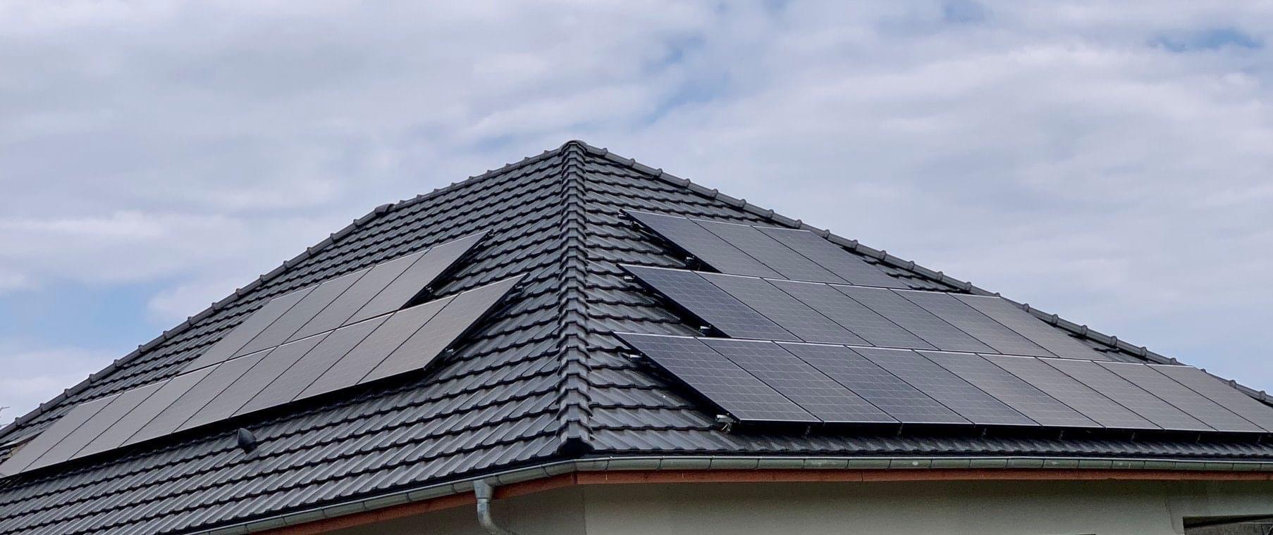 Panele słoneczne na dachu domu pokrytym czarną dachówką ceramiczną, widoczne rynny i fragment ściany budynku, pochmurne niebo w tle.